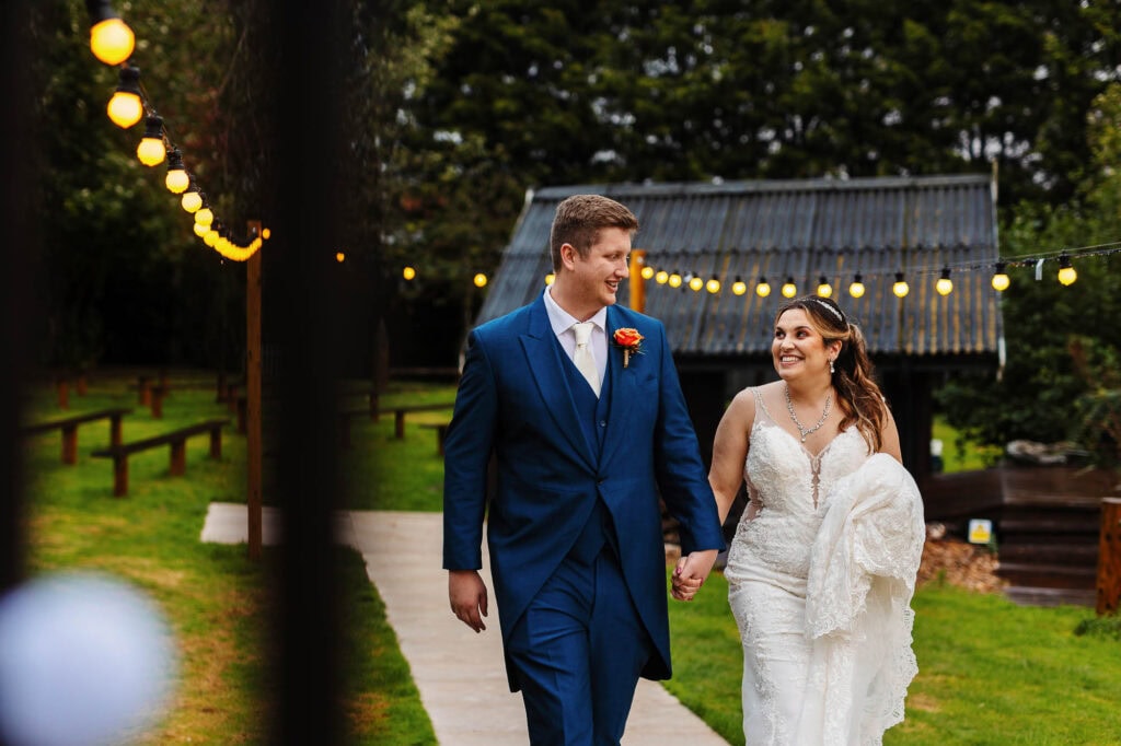 Elegant wedding couple walking hand in hand at outdoor ceremony, romantic Bridal and Groom in wedding attire under string lights, surrounded by lush greenery, celebrating their special day.