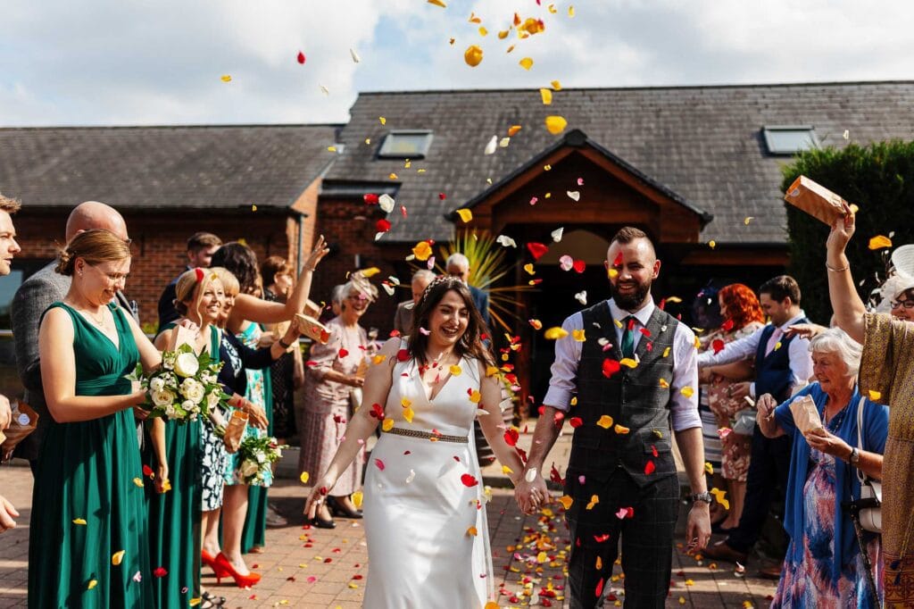 Colourful wedding celebration with guests throwing rose petals at bride and groom outside a rustic barn venue in a sunny day.