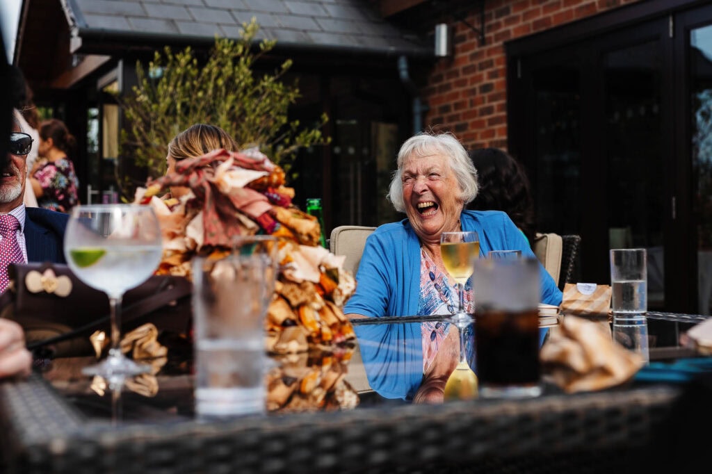 Laughter and joy at a wedding reception outdoor table with guests, drinks, and a delicious group cake, captured by BGS Weddings, showcasing memorable wedding celebration moments.