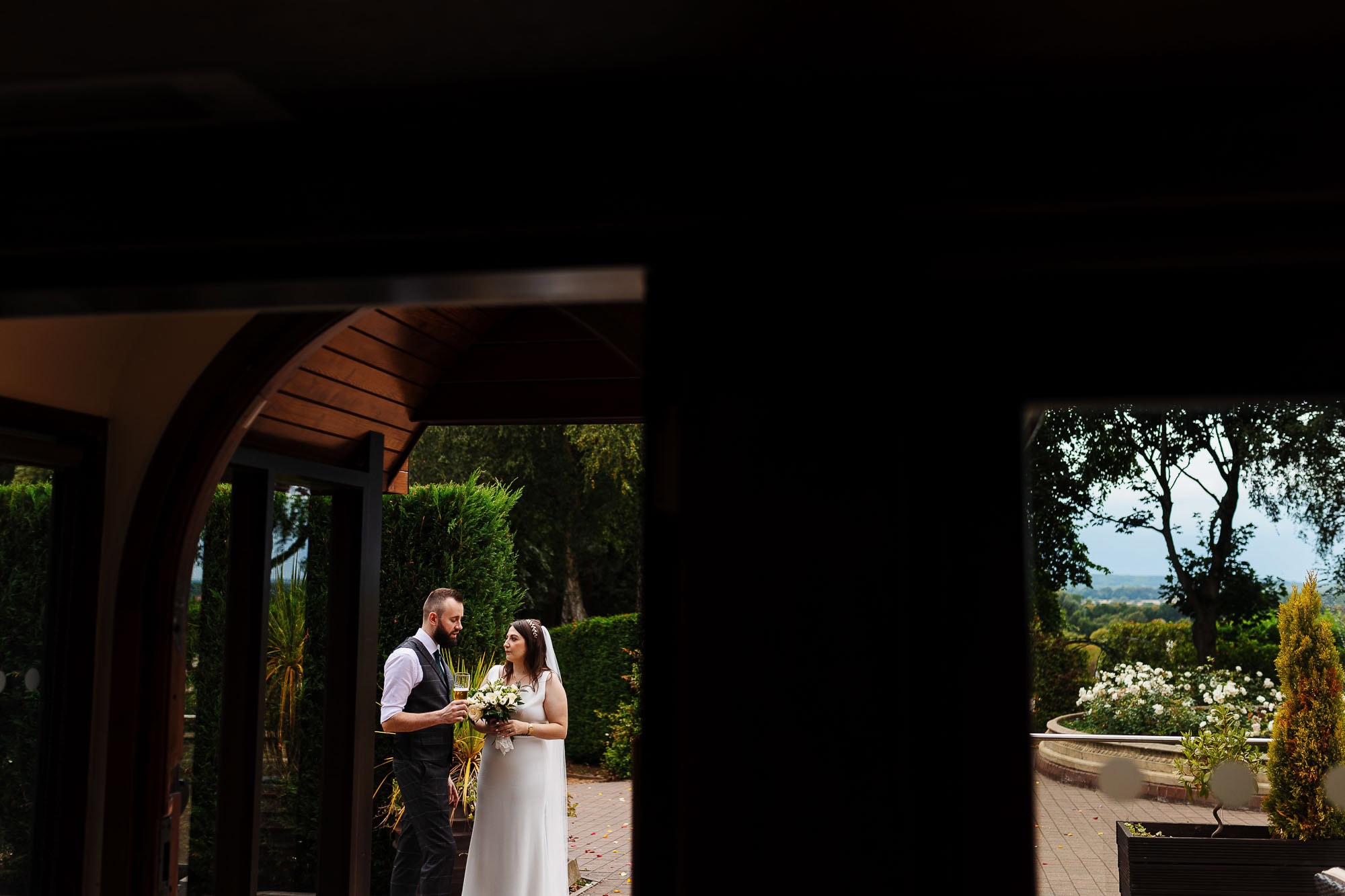 Stunning wedding couple sharing a toast outdoors at a scenic garden venue, captured through an architectural opening, highlighting elegant wedding photography at BGS Weddings, UK.