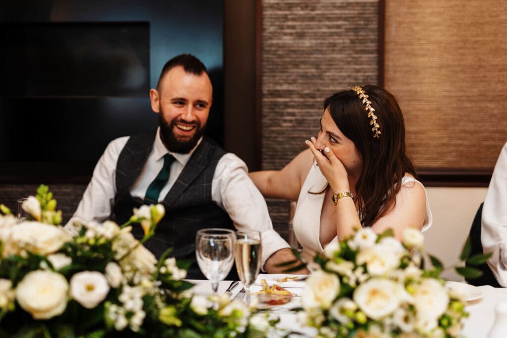 Elegant wedding reception with happy bride and groom sharing a joyful moment at a decorated table with floral arrangements and fine glassware, captured by BGS Weddings.