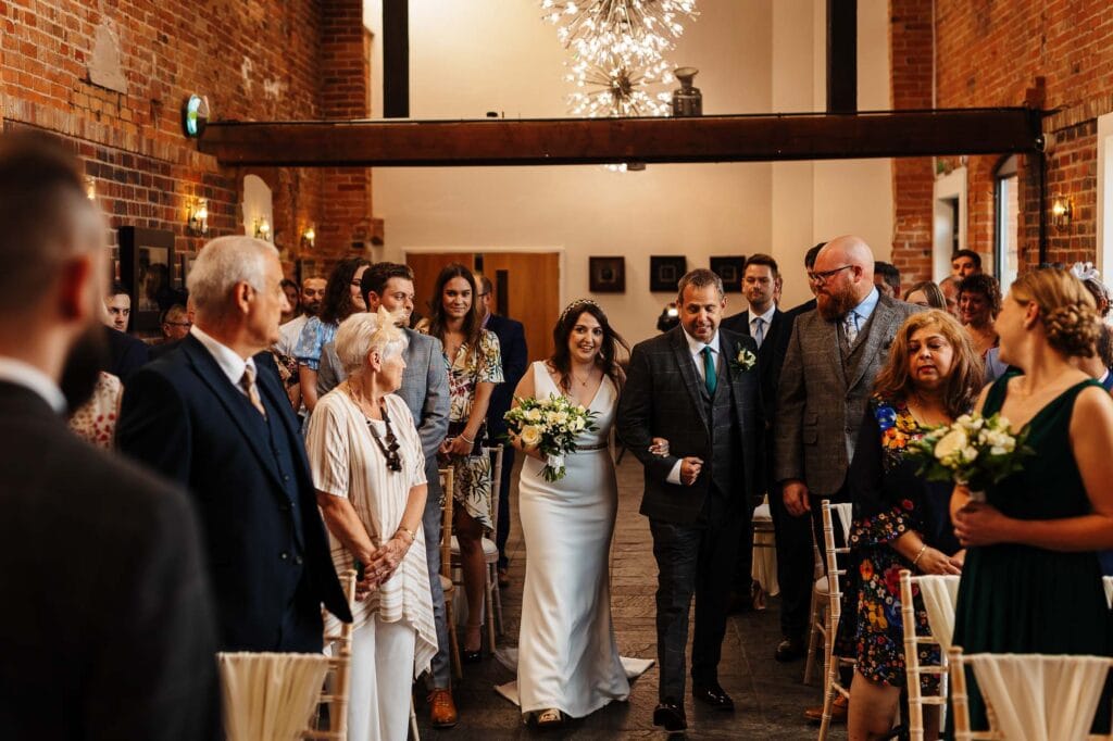 Elegant wedding ceremony featuring the bride walking arm-in-arm with her father in a stylish wedding venue with exposed brick walls and modern ambient lighting.