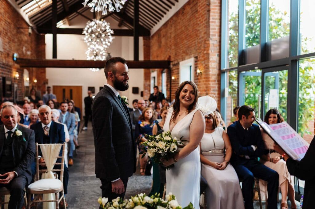 Elegant wedding ceremony at a rustic indoor venue with exposed brick walls and large windows, featuring a happy bride and groom exchanging vows surrounded by guests.