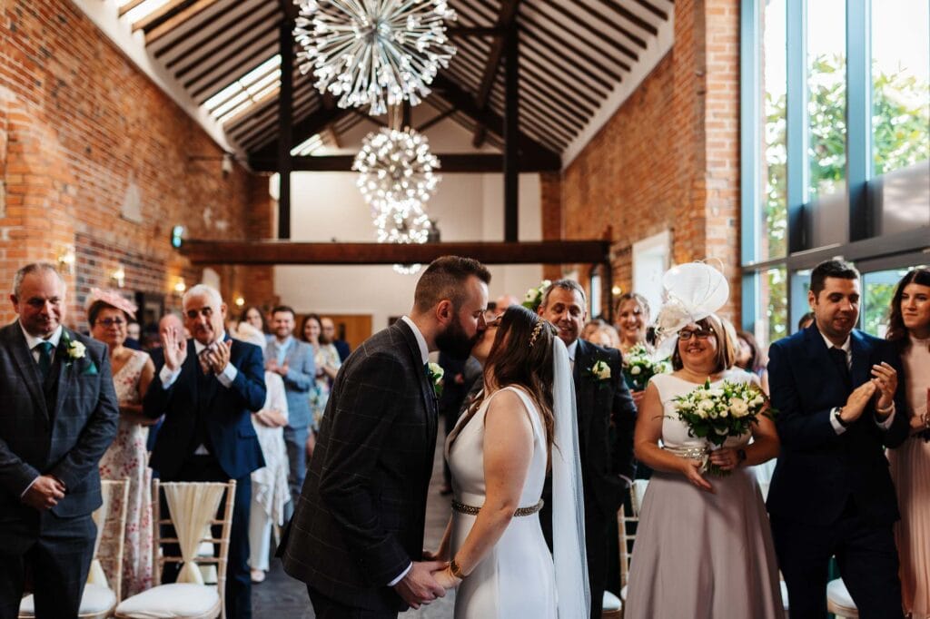 Elegant wedding ceremony at BGS Weddings with bride and groom kissing, surrounded by family and friends in a rustic venue with brick walls and modern chandeliers, celebrating love and marriage.