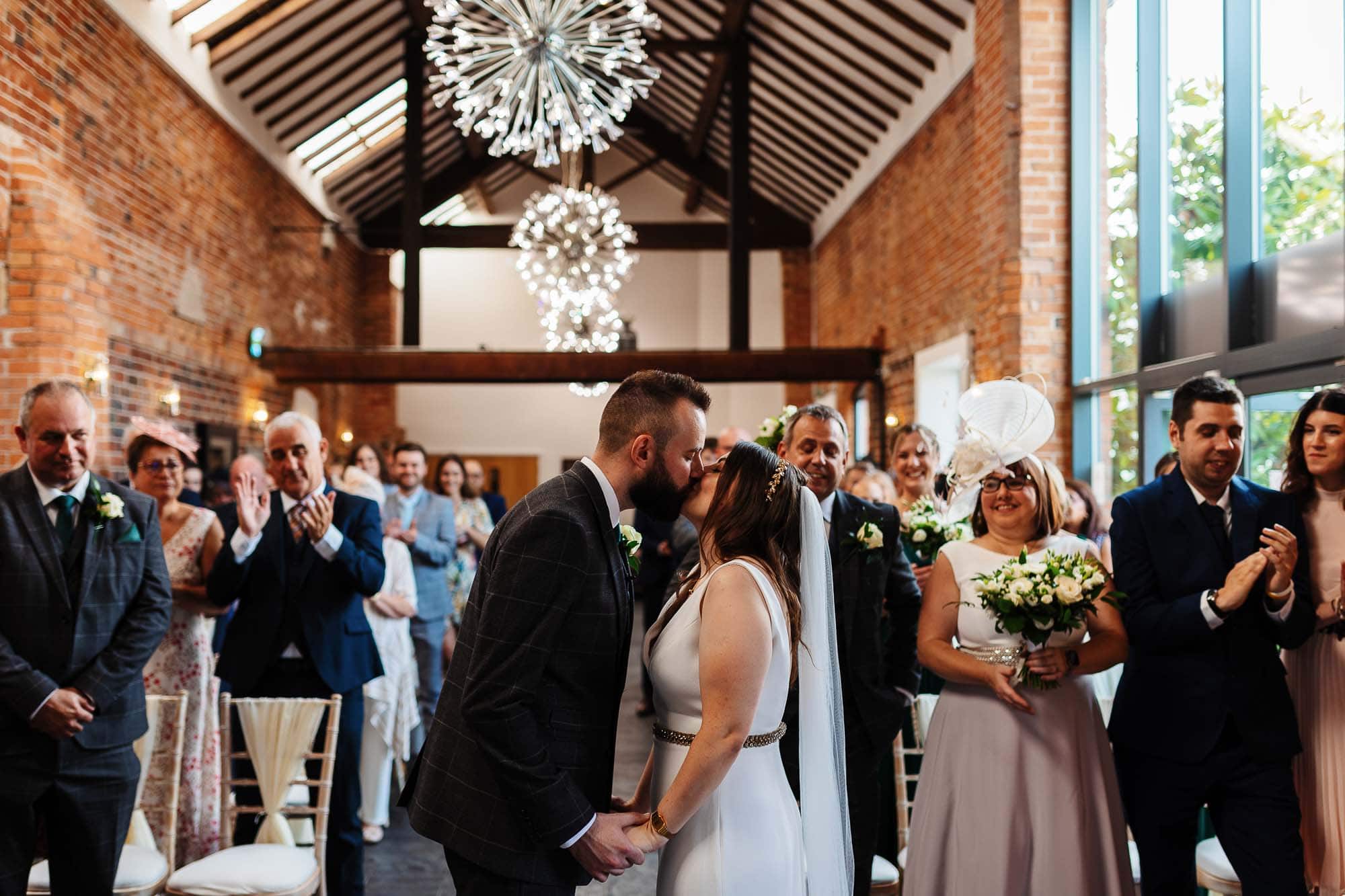 Elegant wedding ceremony at BGS Weddings with bride and groom kissing, surrounded by family and friends in a rustic venue with brick walls and modern chandeliers, celebrating love and marriage.