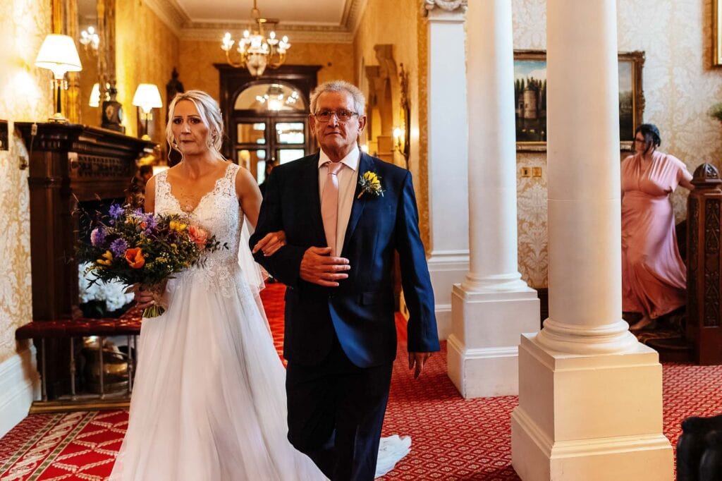 Elegant bride holding a colorful bouquet walking down the aisle after her wedding at BGS Weddings, featuring classic Victorian decor and grand historic architecture.