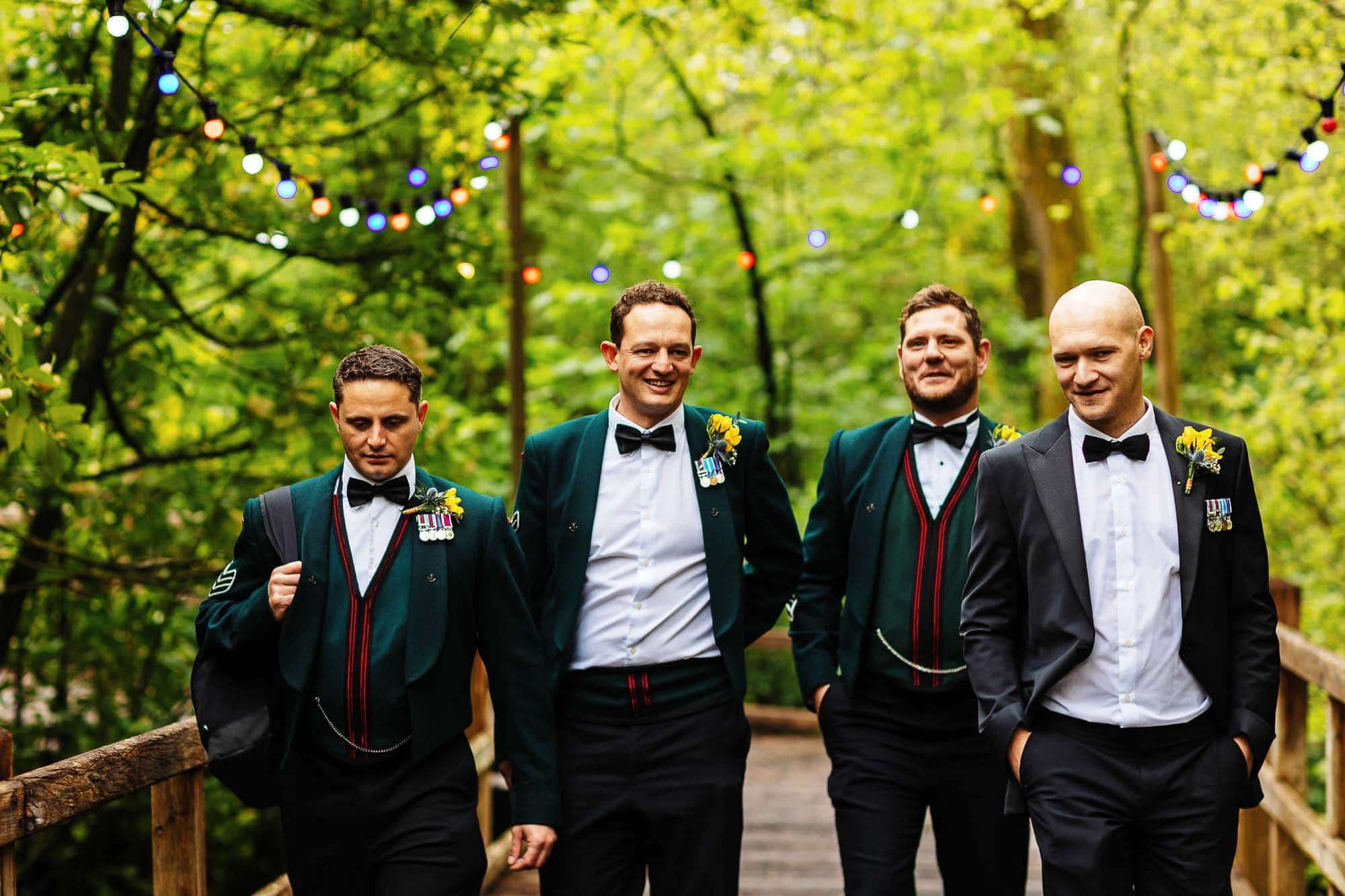 Kilworth House wedding photographer Brightly dressed groomsmen in traditional Scottish kilts and suits walking through a lush green forest with colourful fairy lights overhead at BGS Weddings.