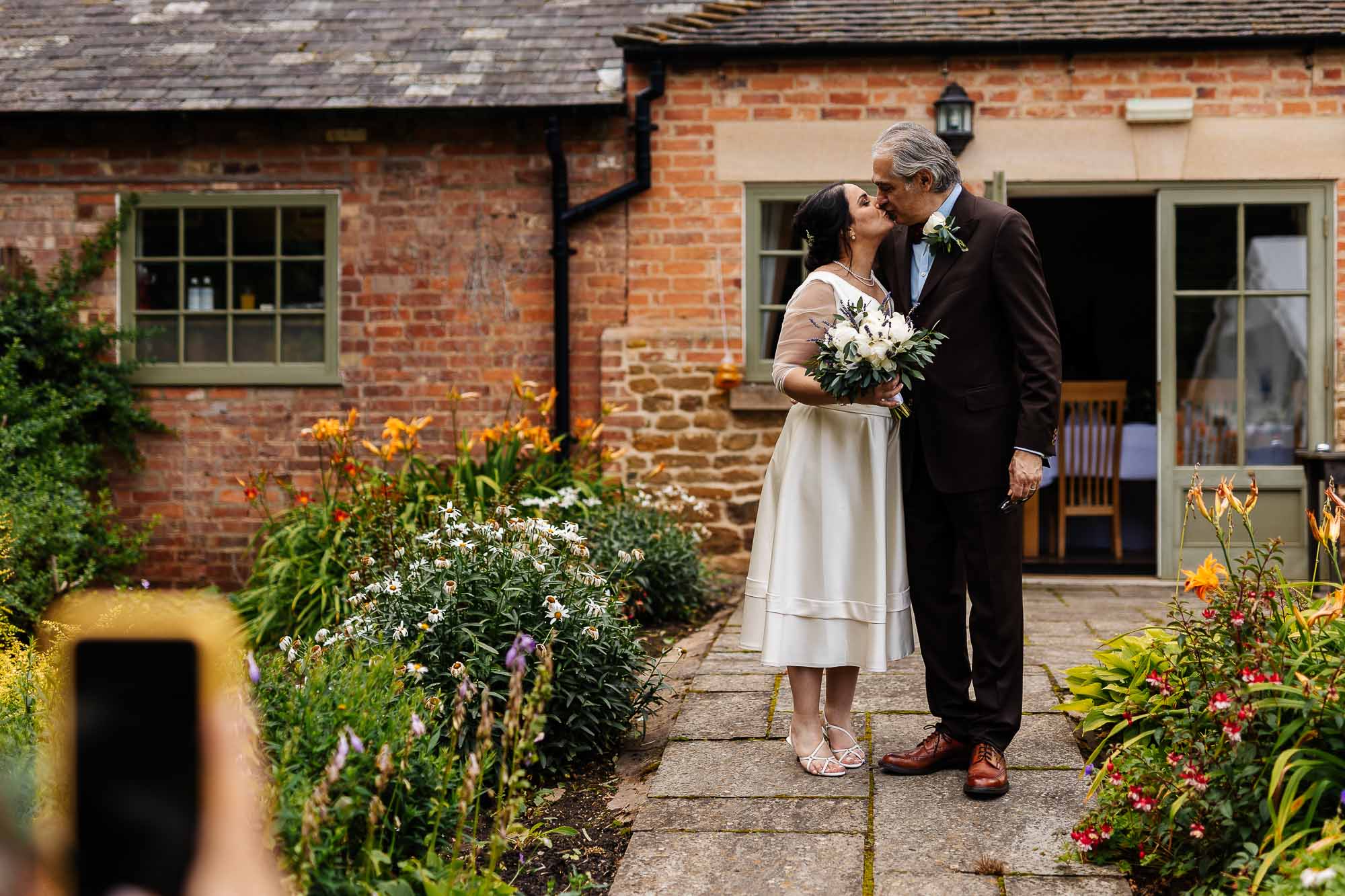 Launde Abbey wedding photographer Elegant wedding couple sharing a kiss outdoors, surrounded by colourful flowers, at a rustic wedding venue by BGS Weddings.