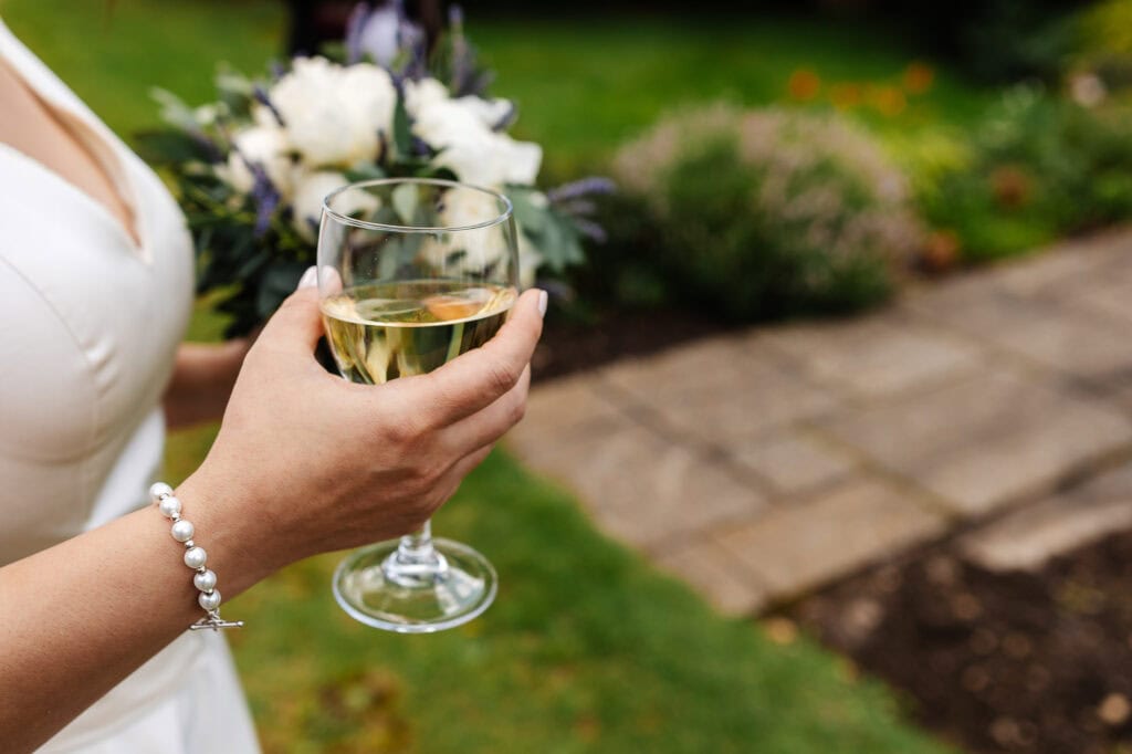 Elegant woman holding a glass of white wine at outdoor wedding celebration with floral arrangements and garden setting.