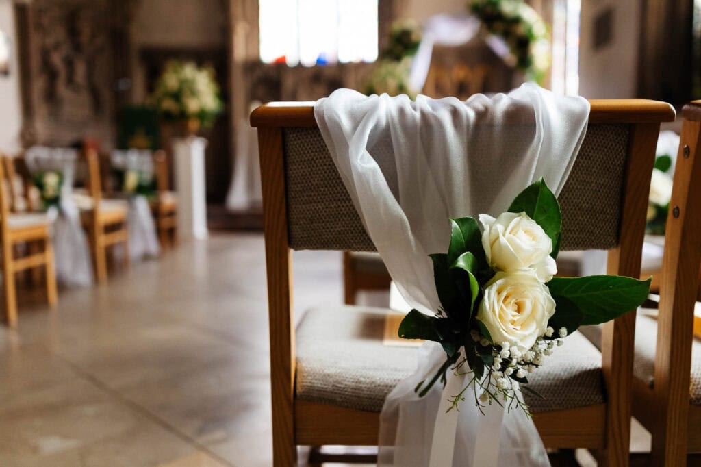 Elegant wedding chair decorated with white roses, greenery, and flowing white tulle, set in a rustic wedding venue with floral arrangements and wooden accents for a romantic ceremony.