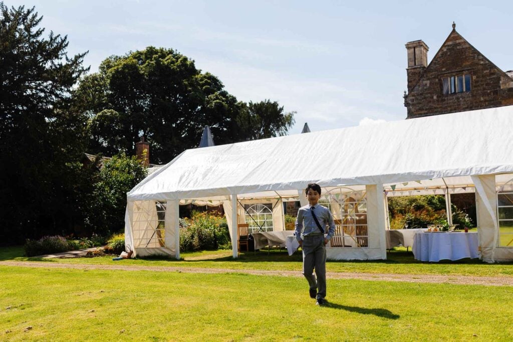 Elegant young boy in formal attire walking across a lush green garden at an outdoor wedding venue, with a large white marquee and historic stone buildings in the background.