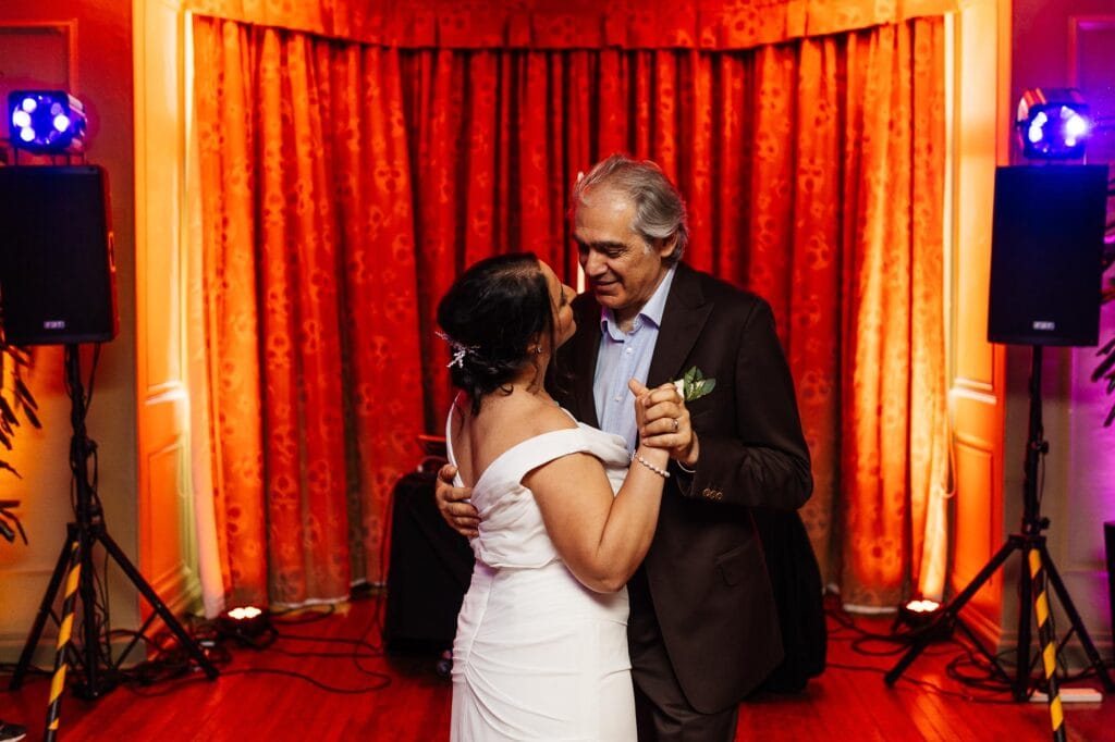 Elegant wedding couple sharing first dance at Bristol wedding venue. Beautifully dressed bride and groom enjoying their special moment with romantic lighting and red curtains.