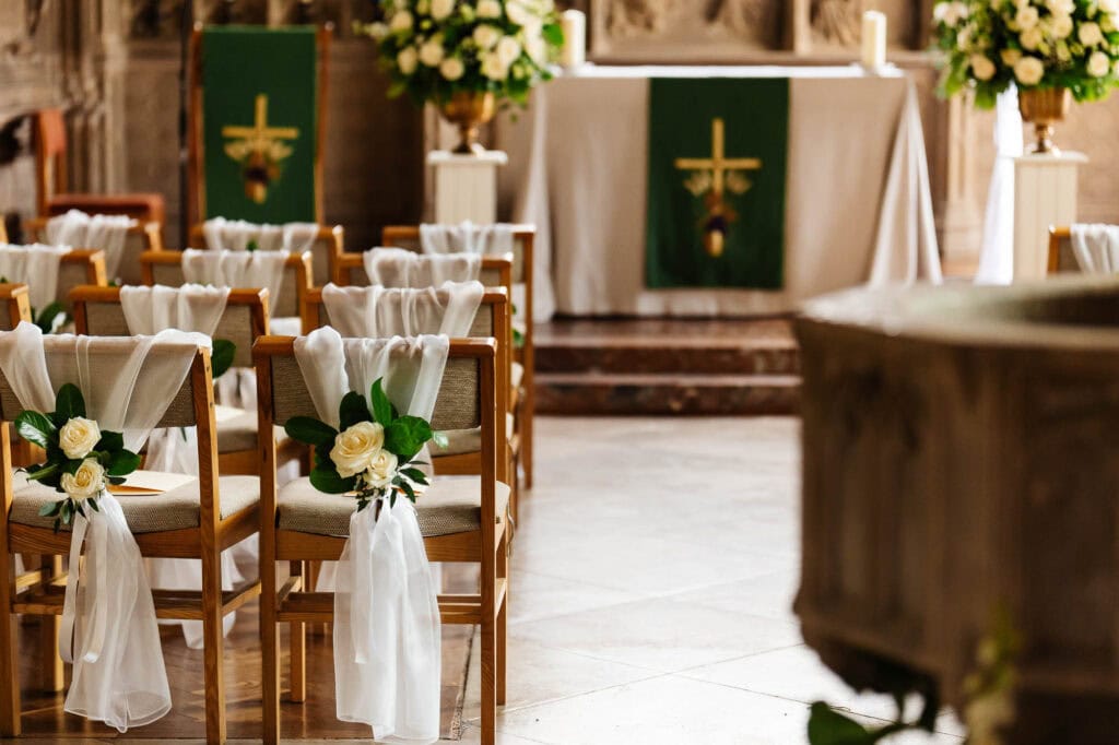 Elegant wedding ceremony chairs decorated with white ribbons and roses in a rustic church setting, ready for a beautiful wedding at BGS Weddings.