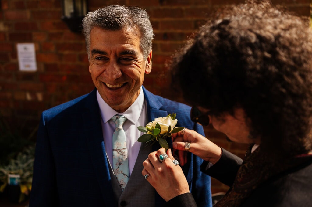 Elegant groom receiving lapel flower pinning at wedding, stylish blue suit, outdoor venue, special occasion, wedding day, beautiful moment, celebrating love, BGS Weddings, UK wedding photography, joyful atmosphere, memorable day.