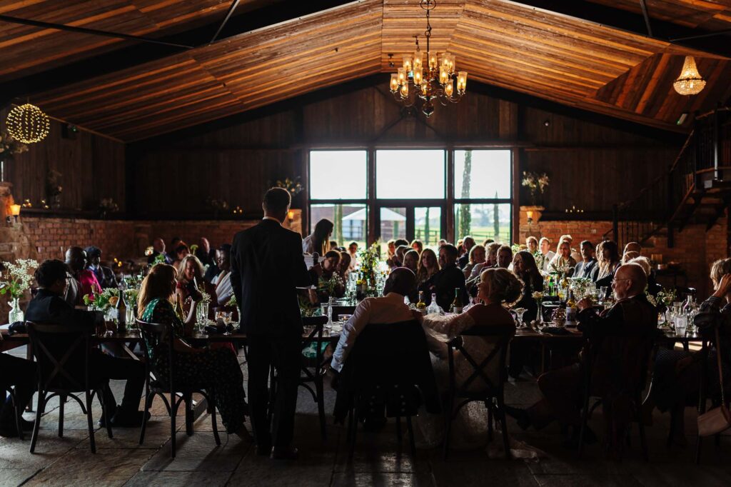 Elegant wedding reception inside a rustic barn with wooden ceilings, chandeliers, and large windows, featuring guests seated at beautifully decorated tables for a romantic wedding celebration.