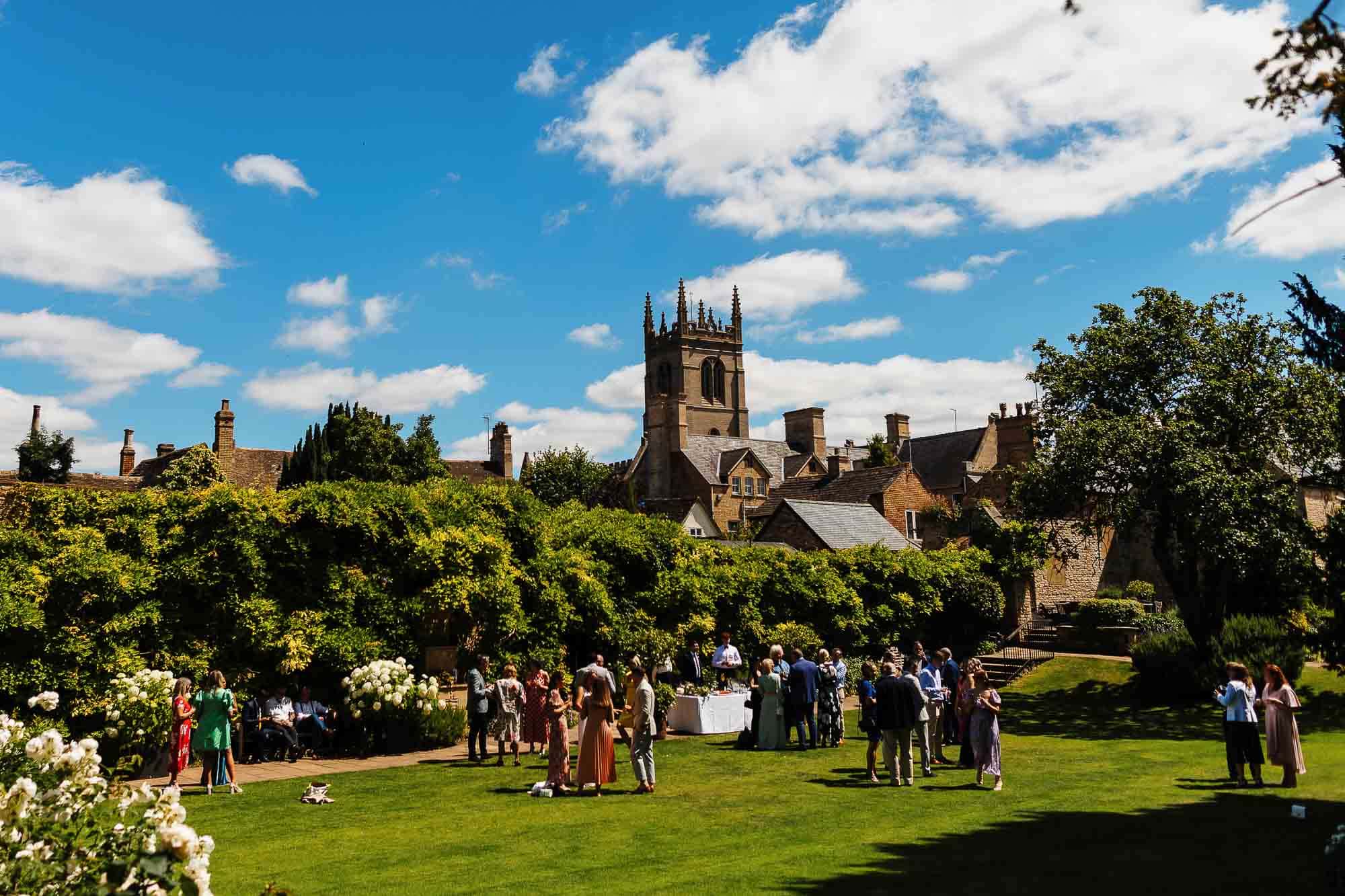 Lincolnshire wedding photographer Elegant outdoor wedding reception with guests socialising on a lush green lawn, historic church in the background, under a bright blue sky, capturing the essence of BGS Weddings in the UK.
