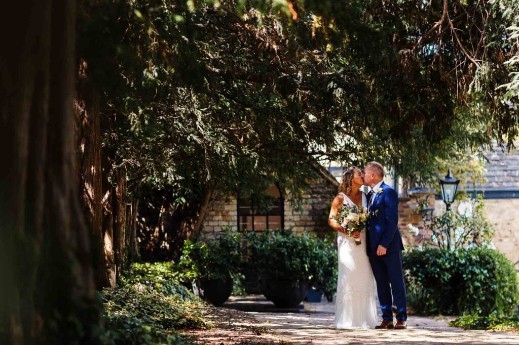 Elegant wedding couple sharing a kiss under a large tree during outdoor wedding ceremony at BGS Weddings, showcasing beautiful wedding photography and romantic atmosphere.