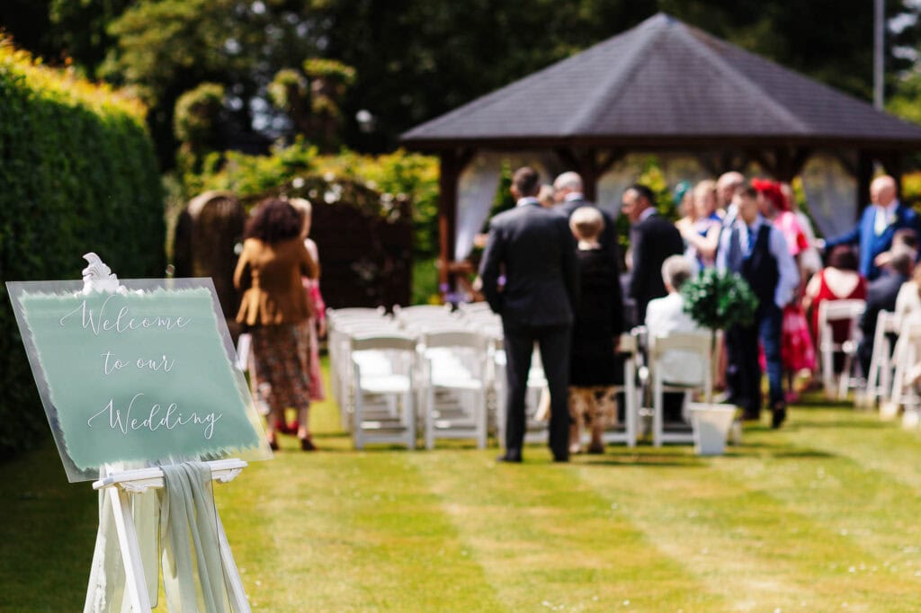 Elegant outdoor wedding ceremony in a lush garden with a welcoming "Welcome to our Wedding" sign, white chairs, and a wooden gazebo, capturing the charm and romance of a perfect wedding day.