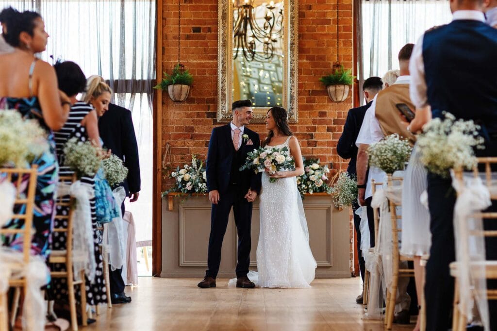 Elegant bride and groom holding hands during their wedding ceremony in a stylish venue with exposed brick walls and floral decor.