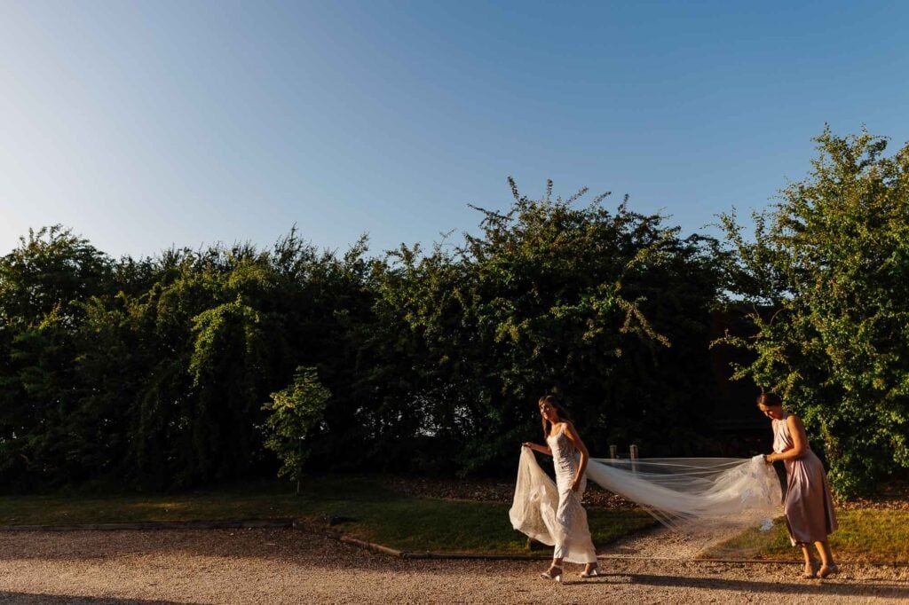 Elegant bride in wedding dress walking on outdoor wedding venue, assisted by bridesmaid holding her veil outdoors during sunset.