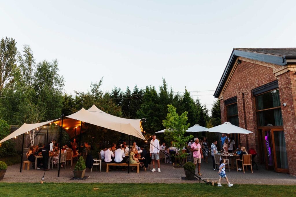 Elegant outdoor wedding reception with string lights, large canopy, and guests enjoying evening celebration in a garden setting.