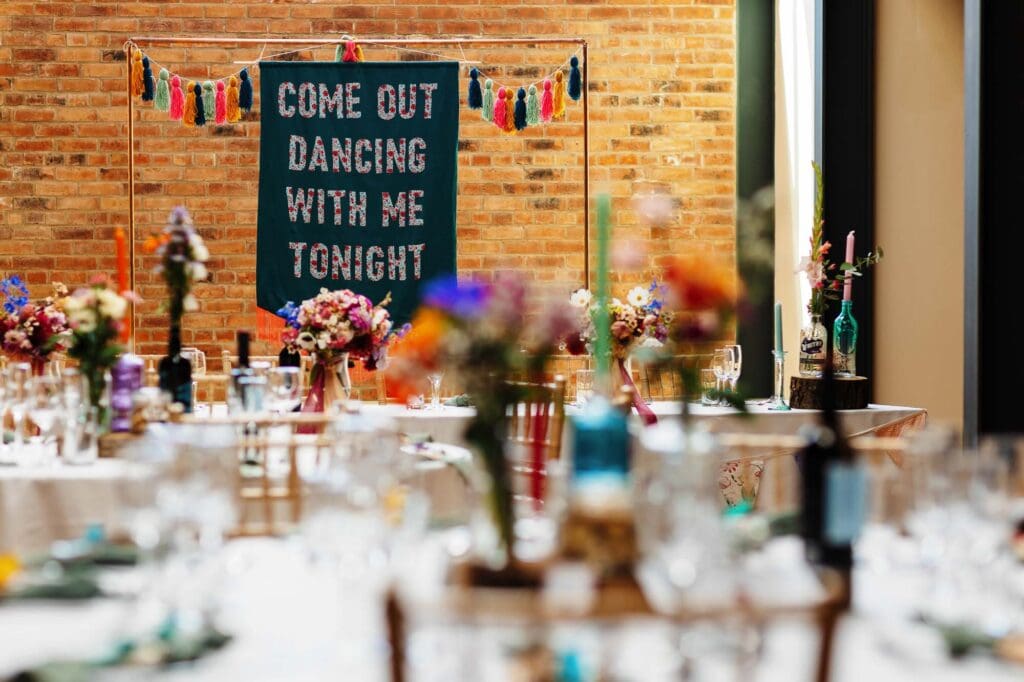 Bright wedding reception table decor at BGS Weddings with colourful floral arrangements and candles, set against a rustic brick wall with a vibrant "Come Out Dancing With Me Tonight" banner, perfect for celebrating love and marriage.