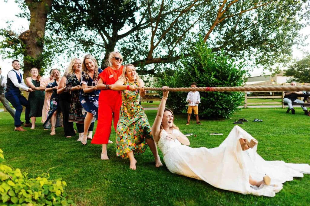 Vibrant outdoor wedding celebration with guests enjoying a spirited game of tug of war in a lush garden setting. The bride and friends are having fun, showcasing a joyful wedding atmosphere.