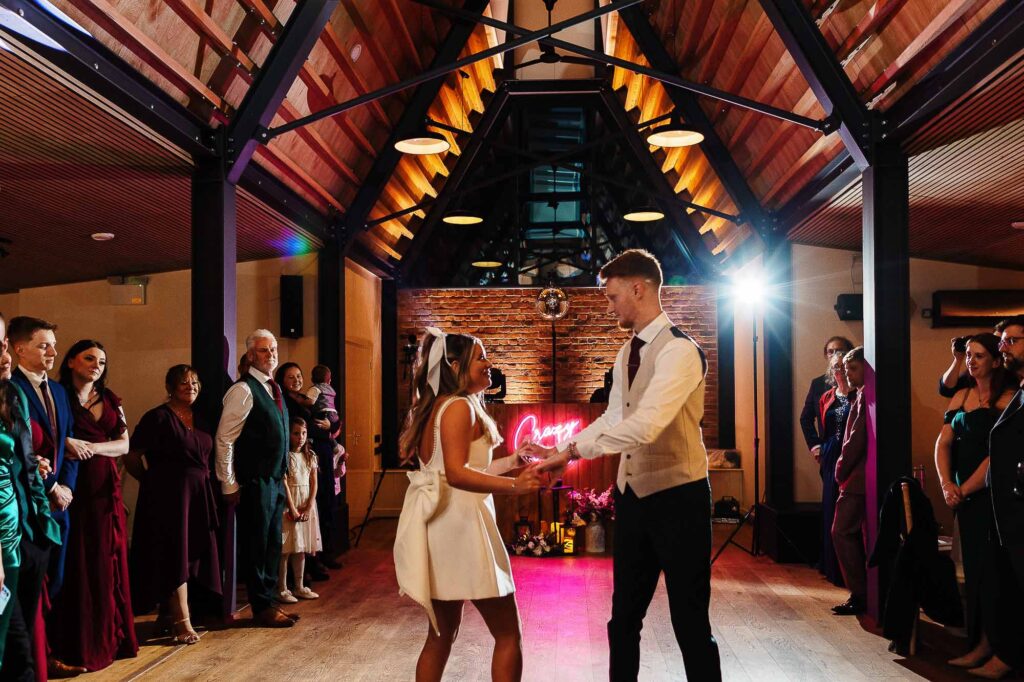 Elegant couple dancing during wedding reception at BGS Weddings venue with guests in formal attire, warm lighting, and rustic brick background, creating perfect wedding celebration atmosphere.