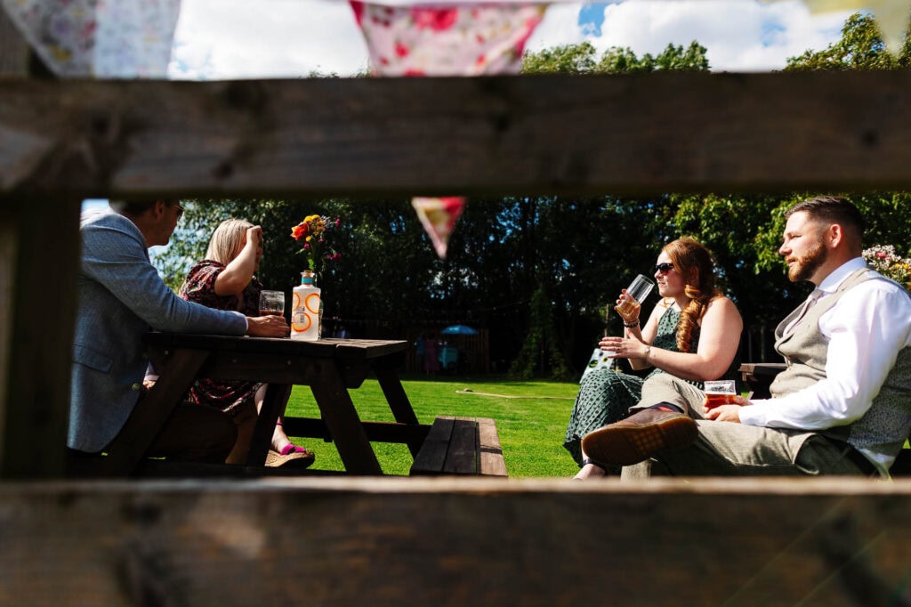 Fun outdoor wedding reception with guests sitting at wooden picnic table, enjoying drinks and conversation on a sunny garden day during a wedding celebration at BGS Weddings.