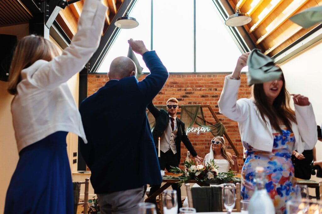 Festive wedding reception with guests dancing, singing and celebrating under a modern glass ceiling at BGS Weddings in Bristol, featuring lively atmosphere and cheerful party vibe.