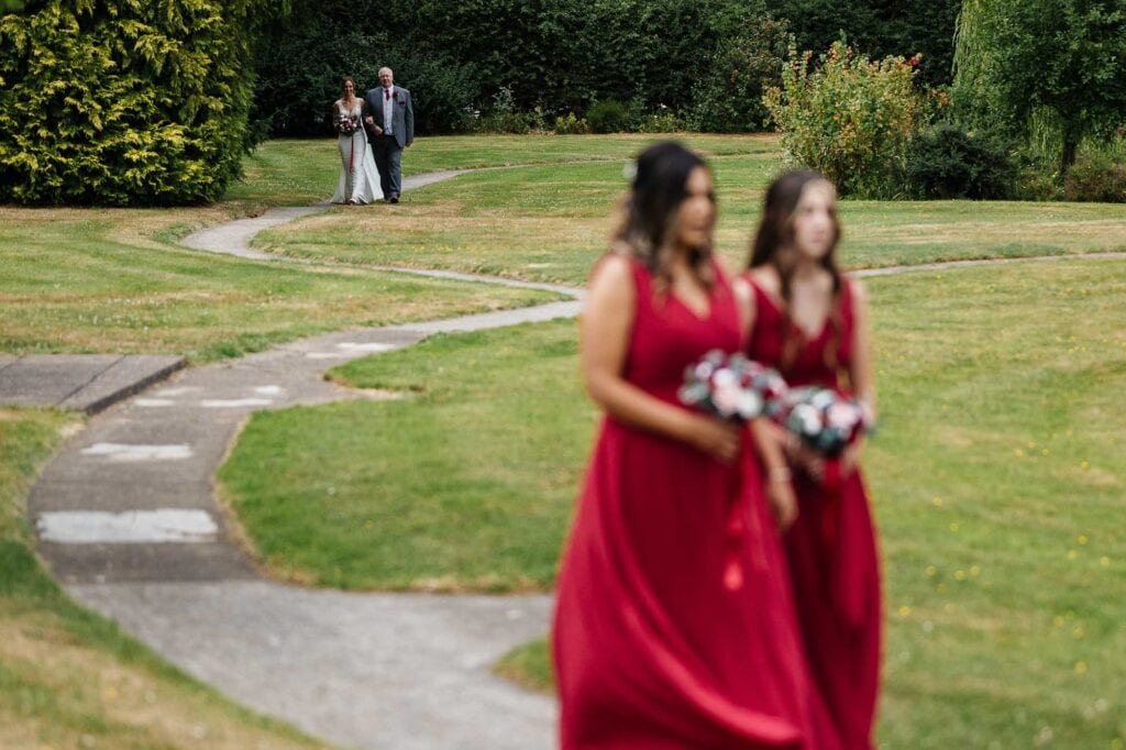 Wedding ceremony processional at BGS Weddings, picturesque outdoor setting with bridesmaids in red dresses and bridal party walking down a landscaped garden pathway.