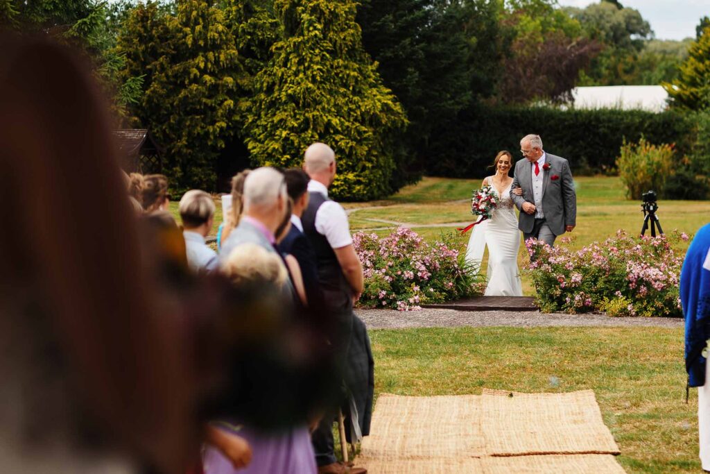 Elegant outdoor wedding ceremony at BGS Weddings with bride walking arm-in-arm with her father among beautiful garden scenery, surrounded by friends and family.