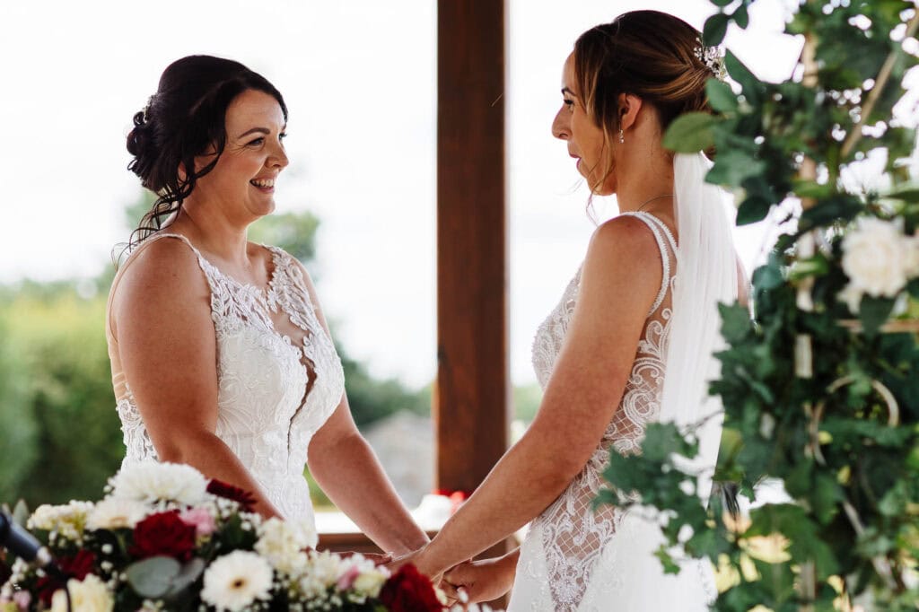 Elegant wedding ceremony with two brides holding hands, wearing beautiful lace wedding dresses, surrounded by floral arrangements, capturing a heartfelt moment at BGS Weddings UK.