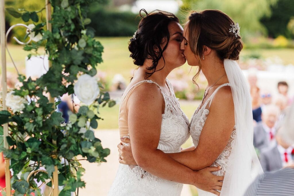 Elegant same-sex wedding kiss between two brides in lace wedding dresses, outdoor ceremony with floral decorations, celebrating love at BGS Weddings.