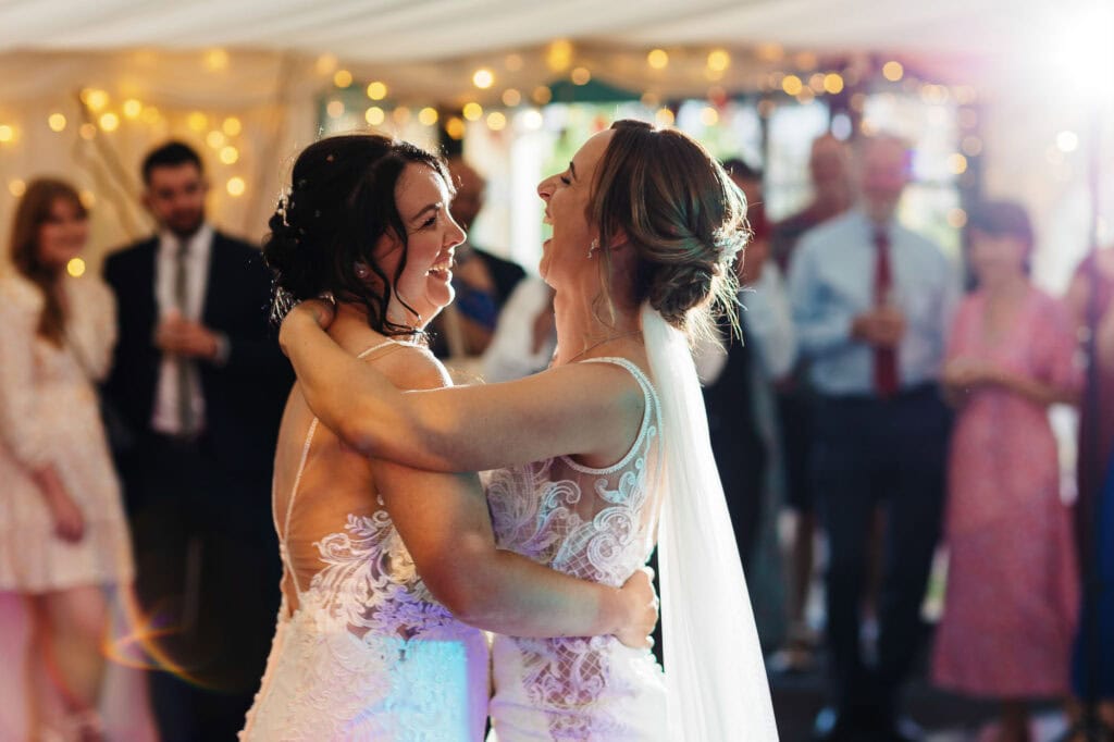 Elegant gay wedding reception with two brides dancing, joyful expression, wedding dress with lace details, indoor celebration, romantic atmosphere, wedding photography, love and happiness, BGS Weddings.