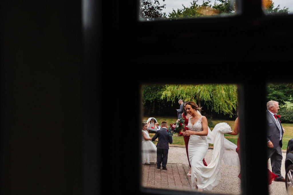 Bride with bouquet arriving at wedding ceremony, smiling bride in a white lace wedding dress, outdoor wedding venue in lush greenery, joyful wedding day moments, BGS Weddings UK.