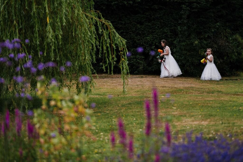 Kids dressed as flower girls in white gowns with floral accessories, holding yellow and pink baskets, walking through a lush garden during a wedding celebration.
