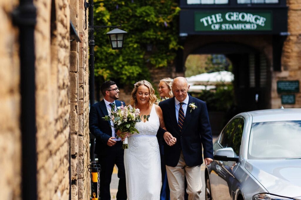 Elegant bride walking with her father, outdoor wedding scene, stylish wedding dress, floral bouquet, Gloucestershire wedding venue, BGS Weddings, romantic wedding celebration in the UK.