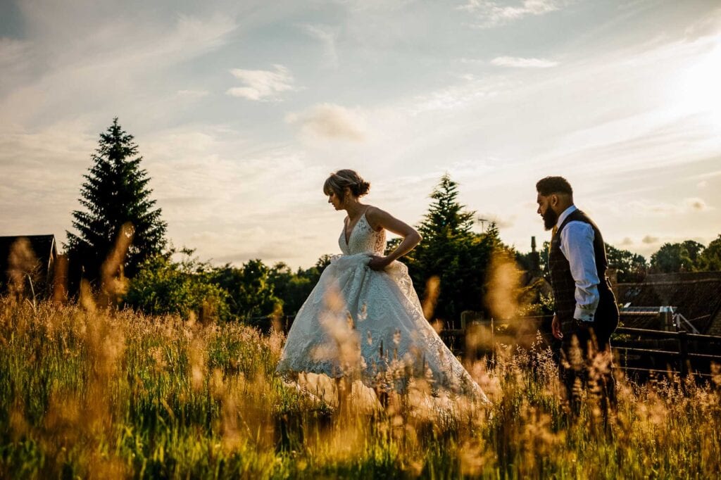 Elegant bride in a lace wedding dress walking through a golden field at sunset, with her groom following, capturing romantic wedding moments outdoors.