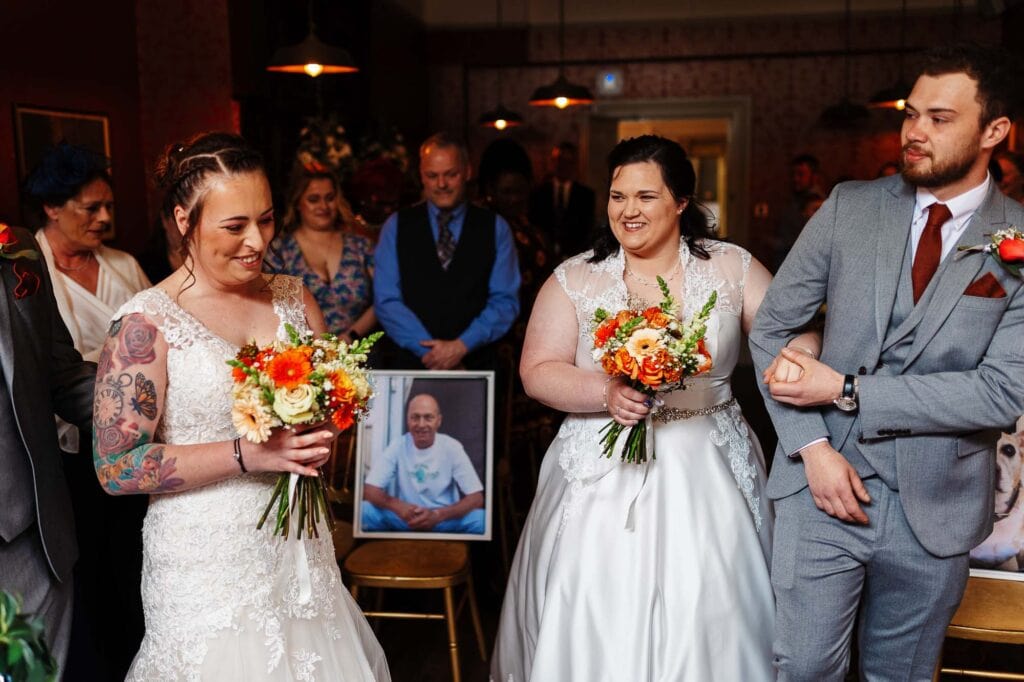 Vibrant wedding scene featuring two brides in white gowns with floral bouquets and a groom in a grey suit, surrounded by friends and family at a formal wedding ceremony.