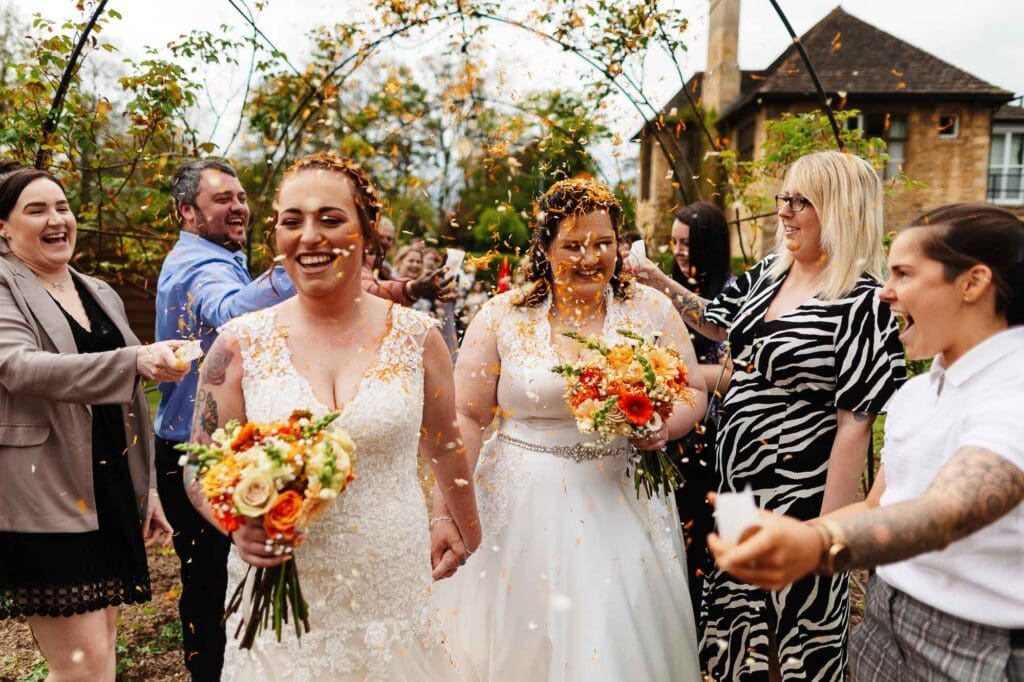 Colorful wedding celebration with two brides holding bouquets, cheering friends, and confetti, outdoors at BGS Weddings, capturing joyful LGBTQ+ wedding moments and real wedding photography in the UK.