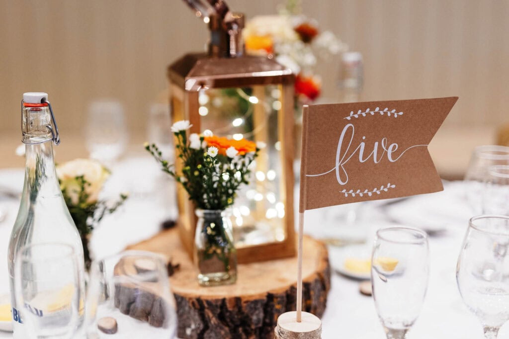 Rustic wedding table centerpiece with a "Five" table number flag, candles, flowers, glass bottles and wood accents at BGS Weddings.