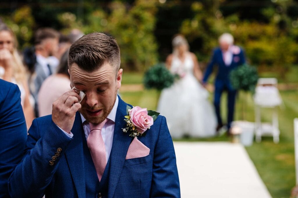 Tearful groom in emotional wedding ceremony outdoors, with bride and guests in background, highlighting heartfelt moments at BGS Weddings, expert wedding photography in the UK.