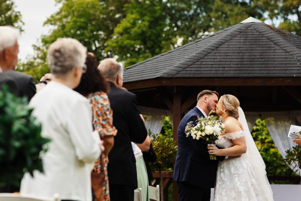 Wedding ceremony at an outdoor gazebo, bride and groom sharing a kiss, surrounded by family and friends, lush greenery in the background, perfect for Richmond wedding venues, professional wedding photography.