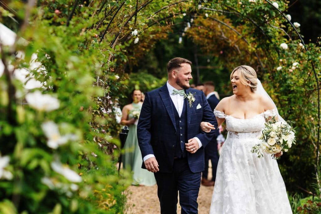 Elegant wedding couple walking under floral archway, celebrating their special day in a picturesque garden setting, with guests in the background.