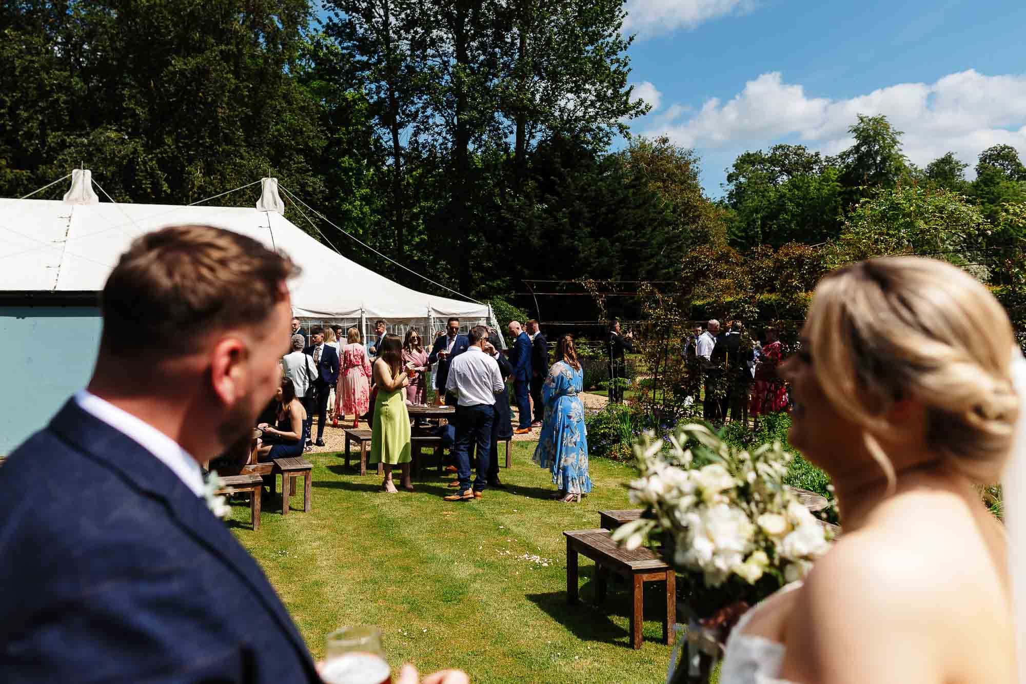 Elegant outdoor wedding reception with guests socialising under a large white marquee in a lush garden setting. Bright, sunny day with vibrant greenery and a festive atmosphere.