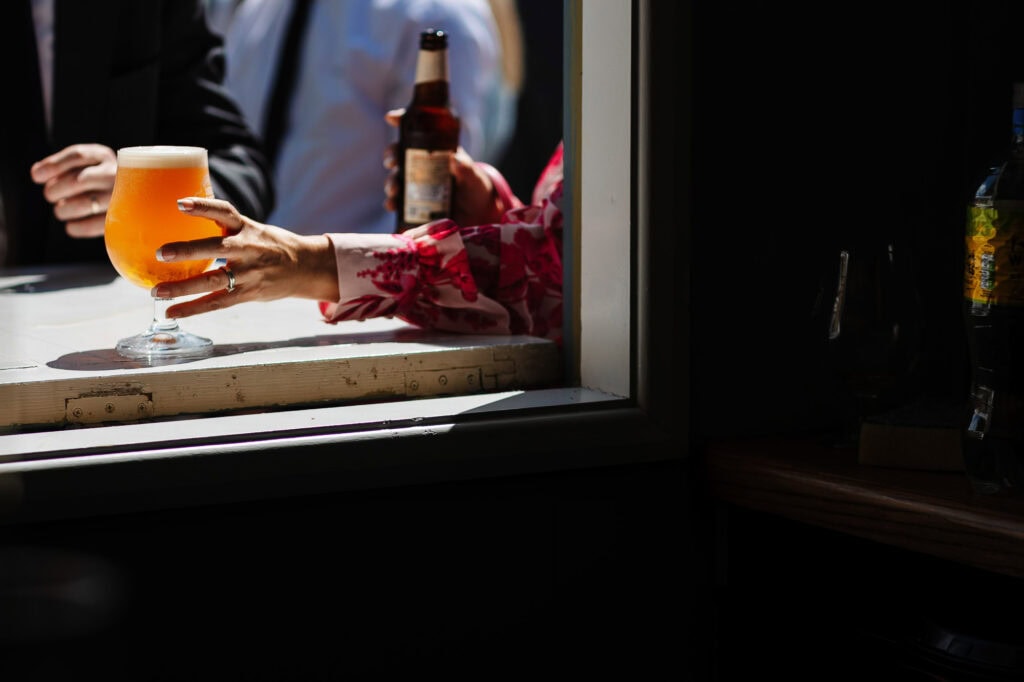 A woman holding a glass of beer and a person with a bottle in the background at a wedding reception, captured through a window with contrasting lighting.