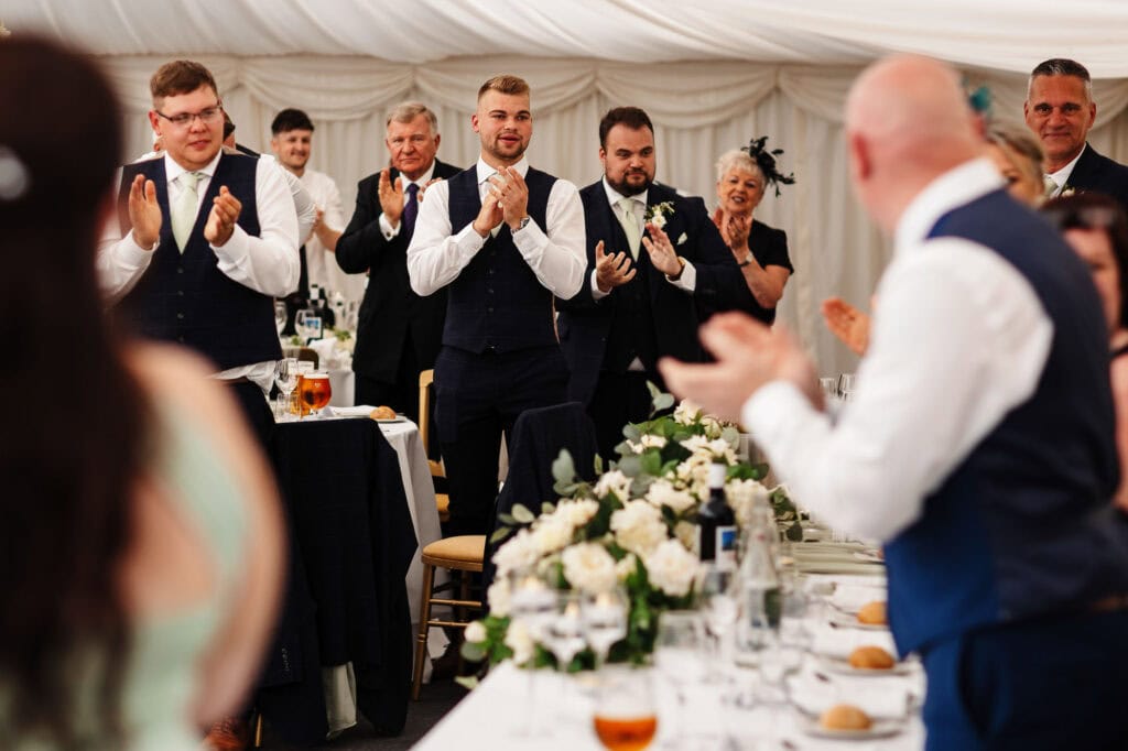 Elegant wedding celebration with guests clapping and toasting in a marquee decorated with floral arrangements, capturing joyful moments from a stylish wedding reception in the UK.