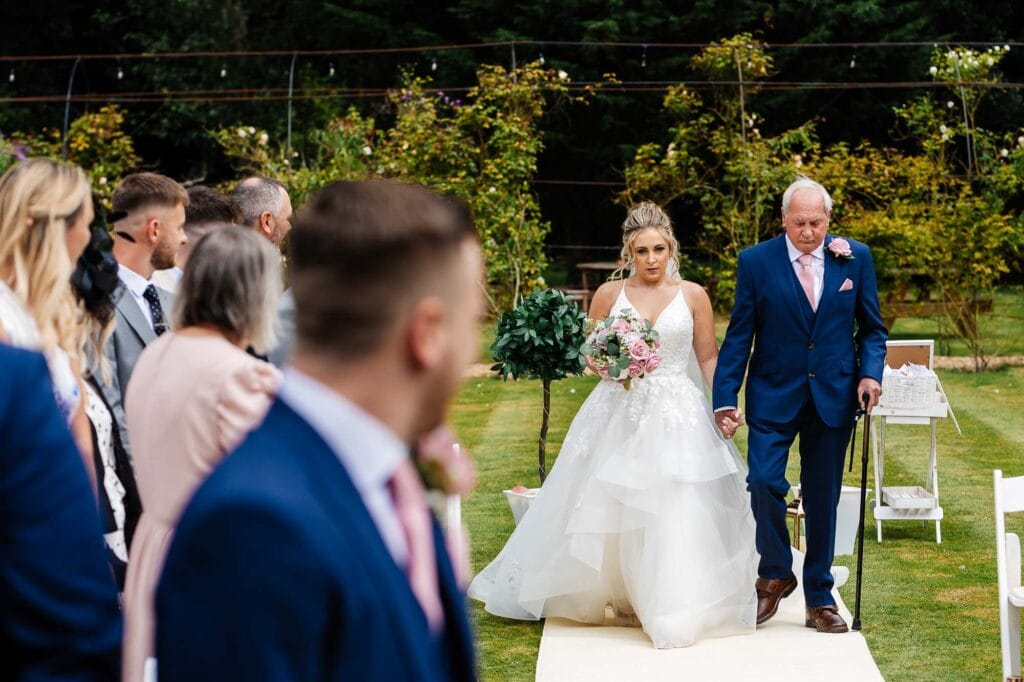 Elegant bride walking down the aisle with her father during outdoor wedding ceremony, surrounded by family and friends, lush green garden in the background.