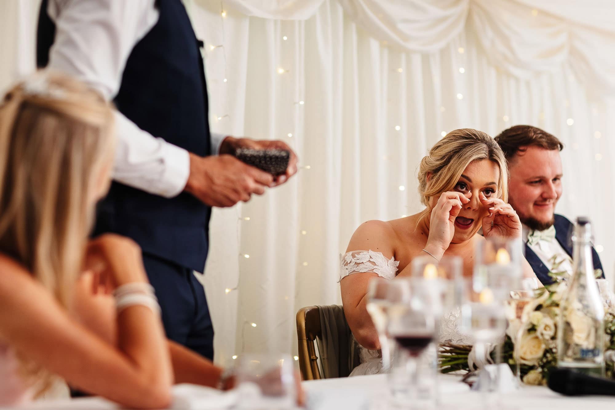 Elegant bride reacting emotionally to speech at wedding reception, beautifully decorated venue with fairy lights, showcasing BGS Weddings' specialised wedding entertainment and event services in the UK.
